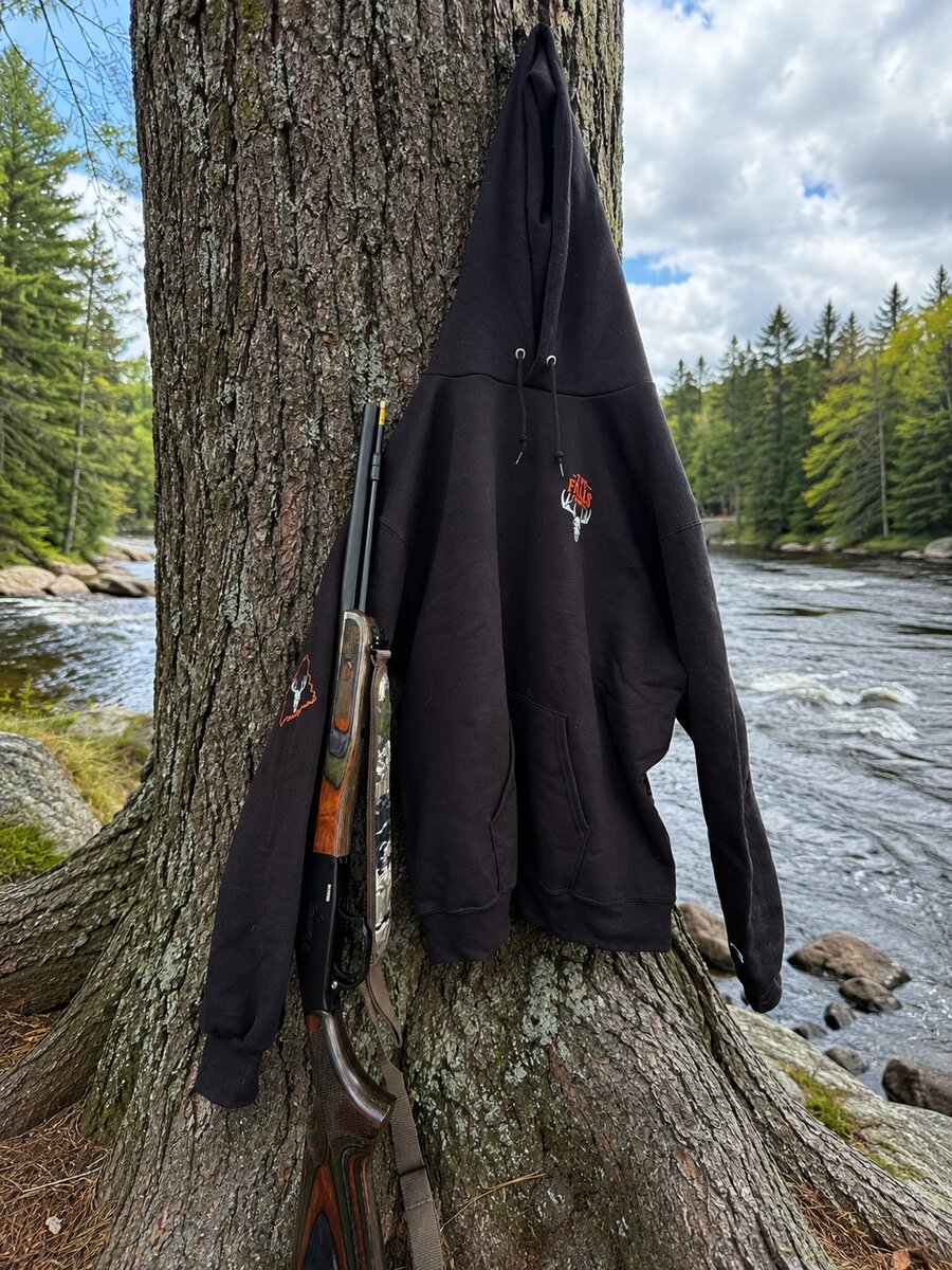 Trails End hoodie hanging on a tree beside a rifle, next to the river